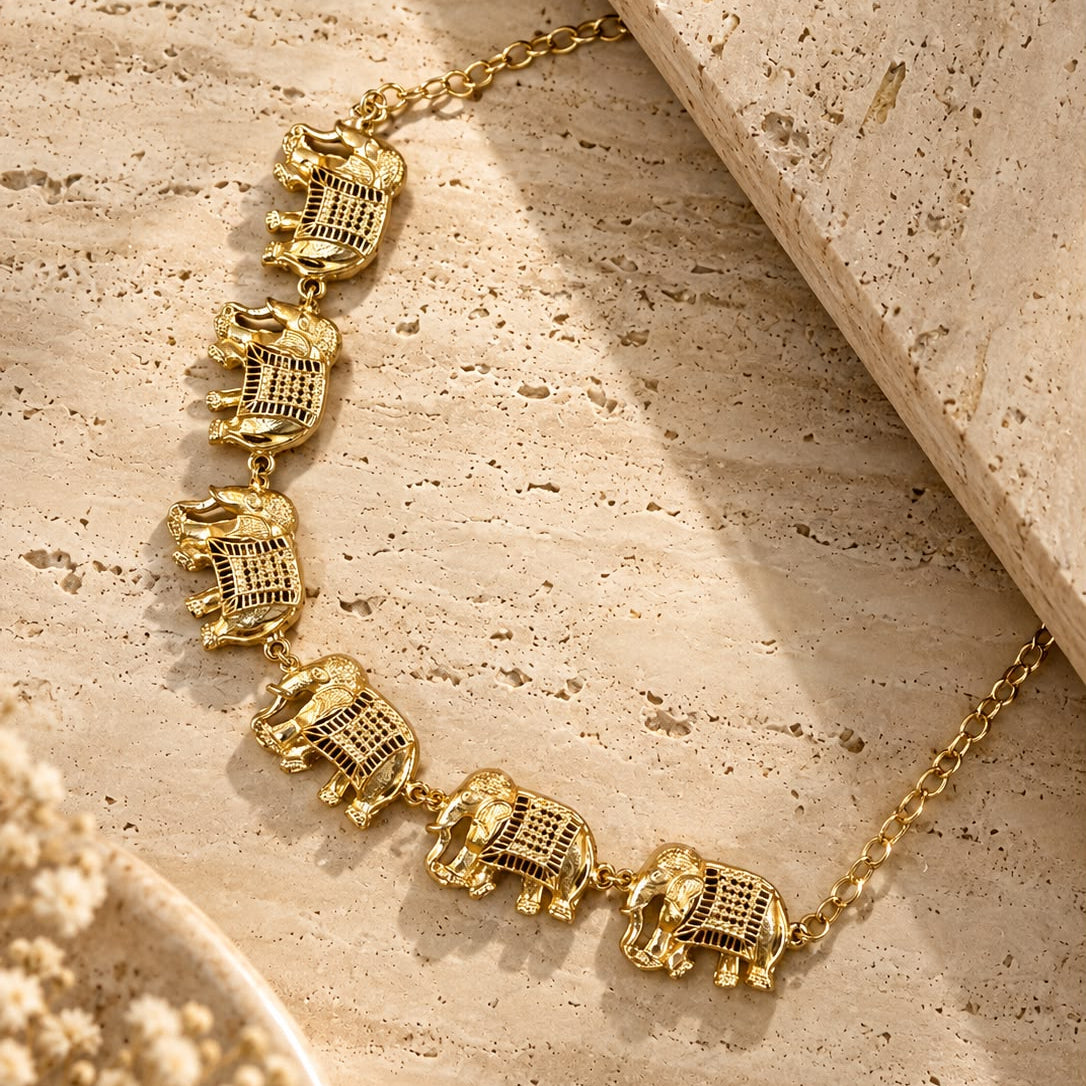 A gold-toned elephant necklace with textured details, displayed on a beige travertine surface with dried flowers in the corner.