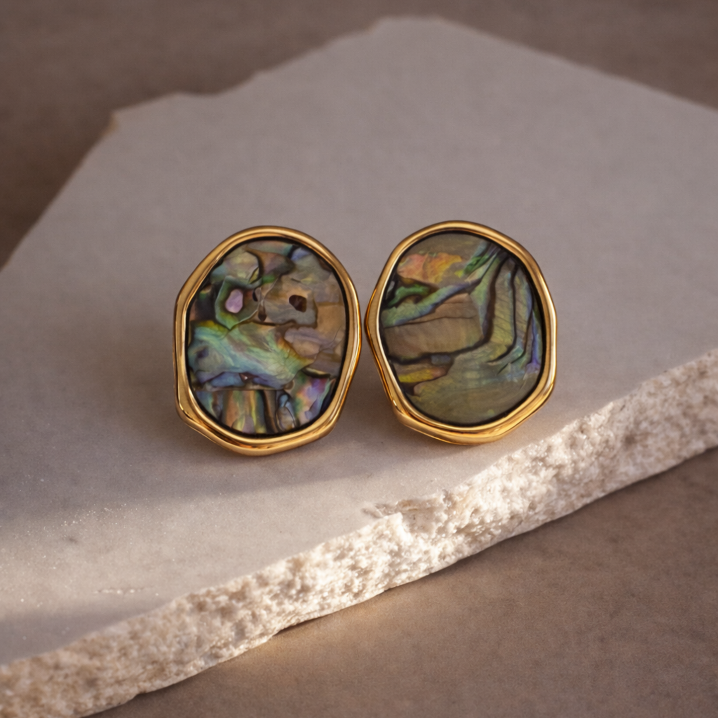 Pair of gold-framed holographic shell earrings on a stone surface