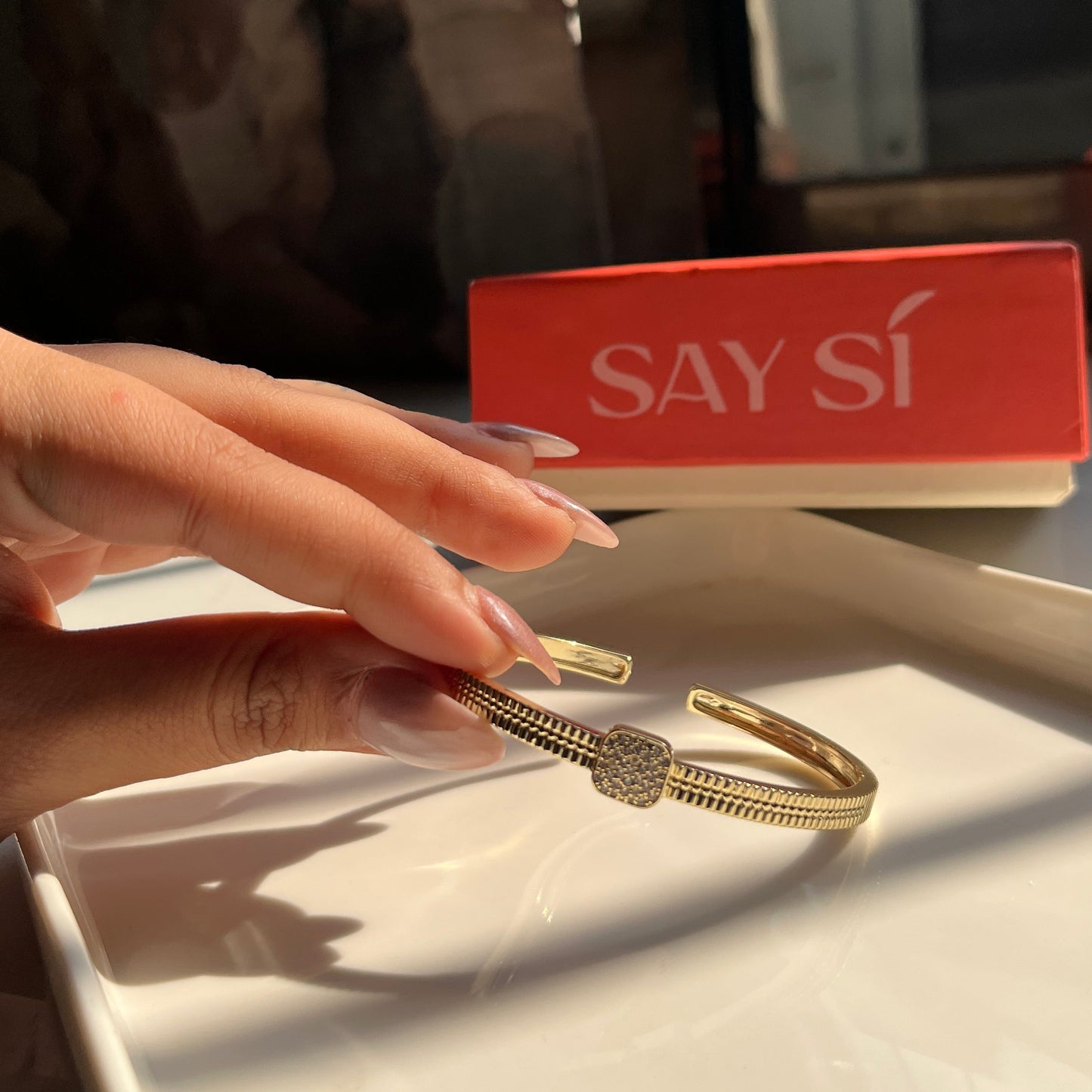 Hand holding a gold bracelet with a Say Si jewellery box in the background