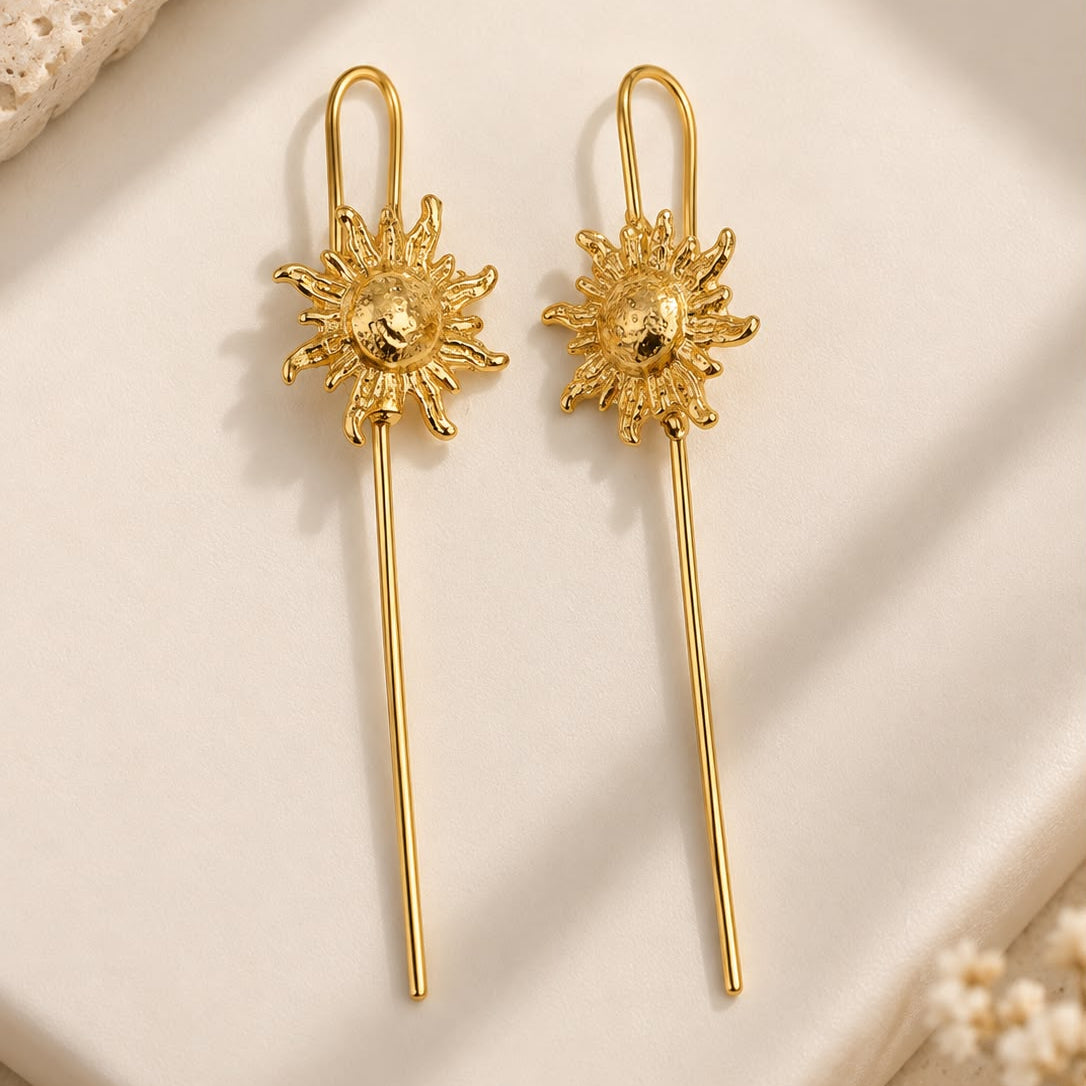 A pair of gold-toned long dangle earrings with textured sunburst charms, displayed on a cream-colored ceramic tile with neutral decorative props.