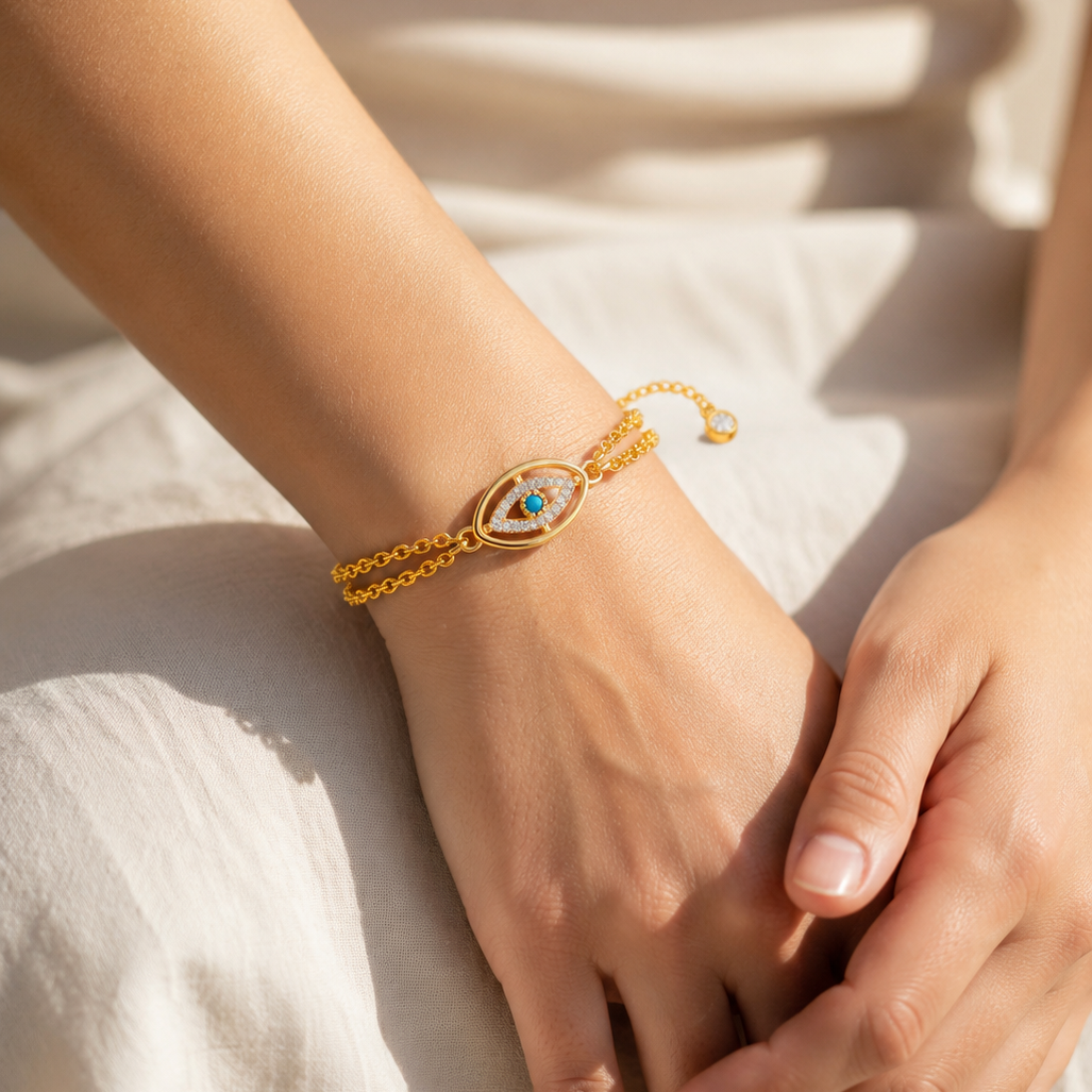 Gold bracelet with a blue evil eye stone on a person's wrist, wearing a cream outfit.