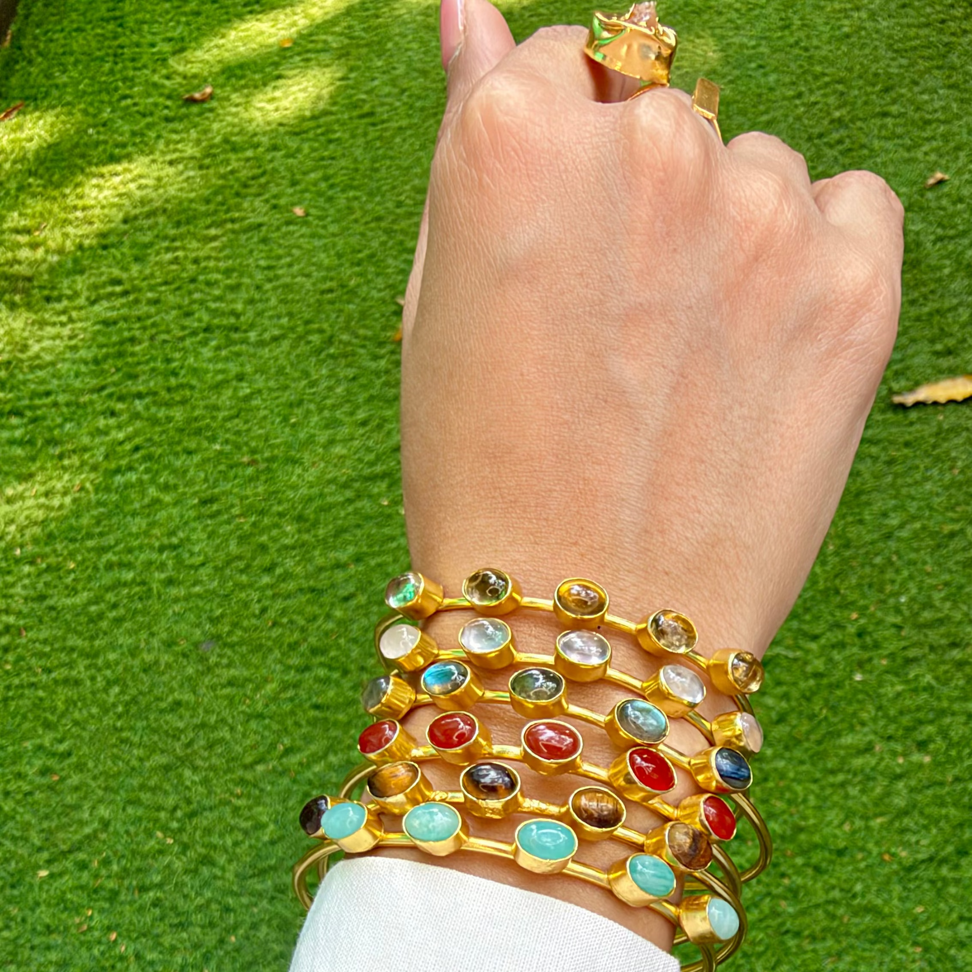 A close-up of a person's wrist wearing a stack of gold-plated bangles adorned with colorful round and oval gemstones, set against a green grass background.
