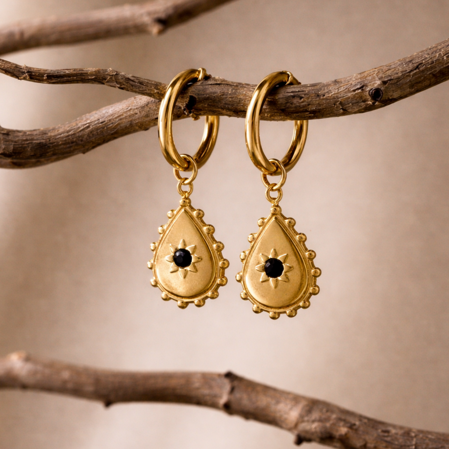 Noir droplet hoop earrings, dangling on a branch against a beige background