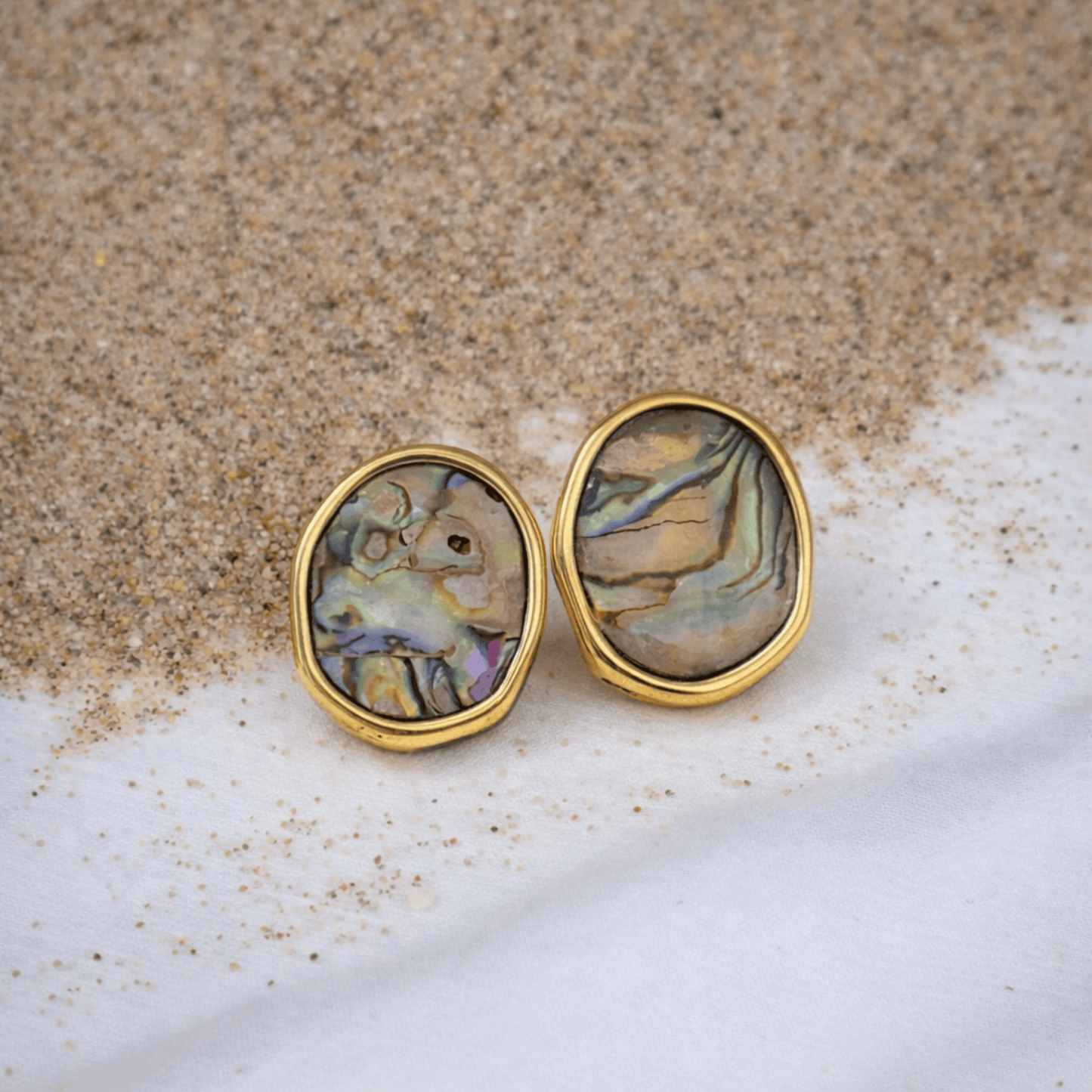 Pair of oval earrings with multicolored holographic stone centers on a sandy surface
